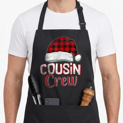 Cousin christmas plaid apron - Red and black plaid Santa hat above white COUSIN Crew text with distressed texture — shown on a black cotton canvas apron against a white background
