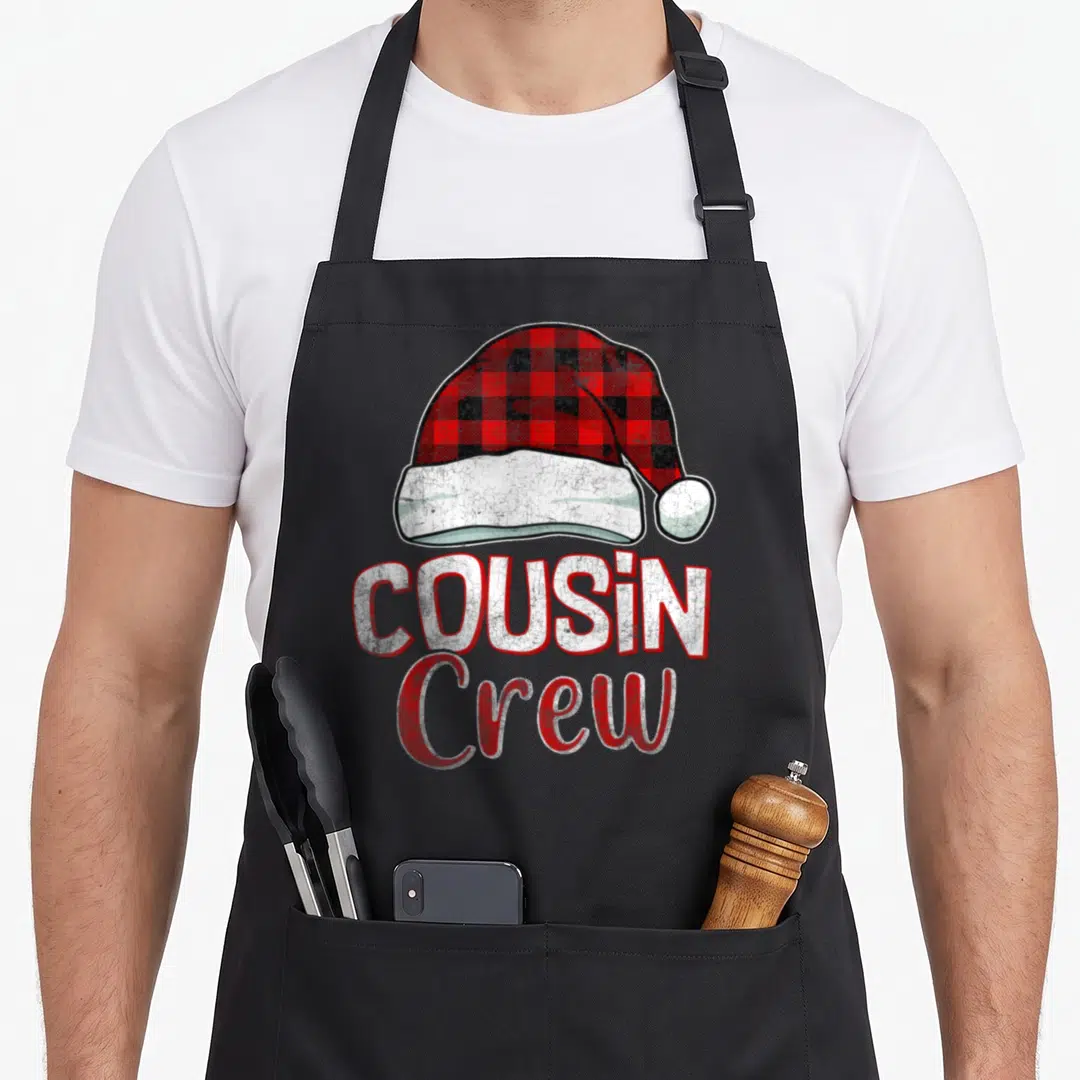 Cousin christmas plaid apron - Red and black plaid Santa hat above white COUSIN Crew text with distressed texture — shown on a black cotton canvas apron against a white background