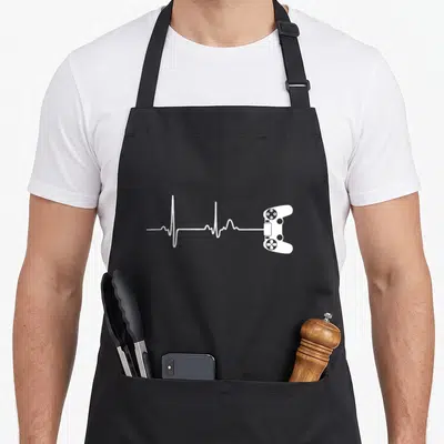 Player video game apron - White heartbeat line connecting to a game controller outline on black fabric — shown on a black cotton canvas apron against a white background