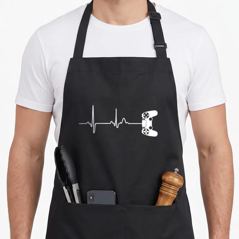 Player video game apron - White heartbeat line connecting to a game controller outline on black fabric — shown on a black cotton canvas apron against a white background