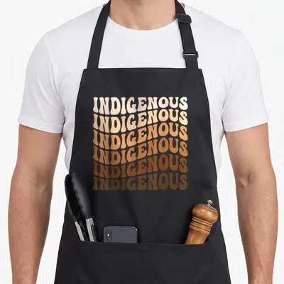 Heritage indigenous native american aprons - Stacked INDIGENOUS text in retro font with cream to brown gradient — shown on a black cotton canvas apron against a white background