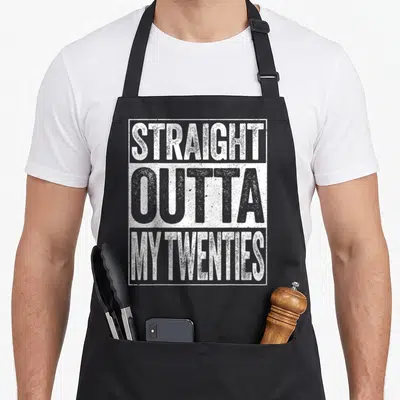 Straight birthday apron - Stacked white and grey distressed typography reading STRAIGHT OUTTA MY TWENTIES inside — shown on a black cotton canvas apron against a white background