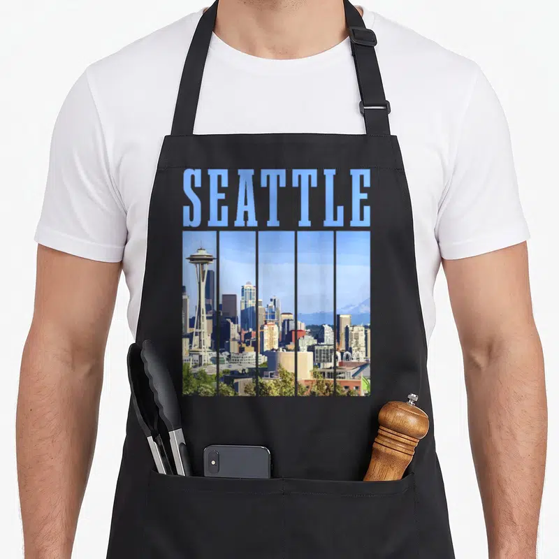 Pride seattle apron - SEATTLE text above retro skyline split into vertical panels with grey stripes and Space Needle — shown on a black cotton canvas apron against a white background