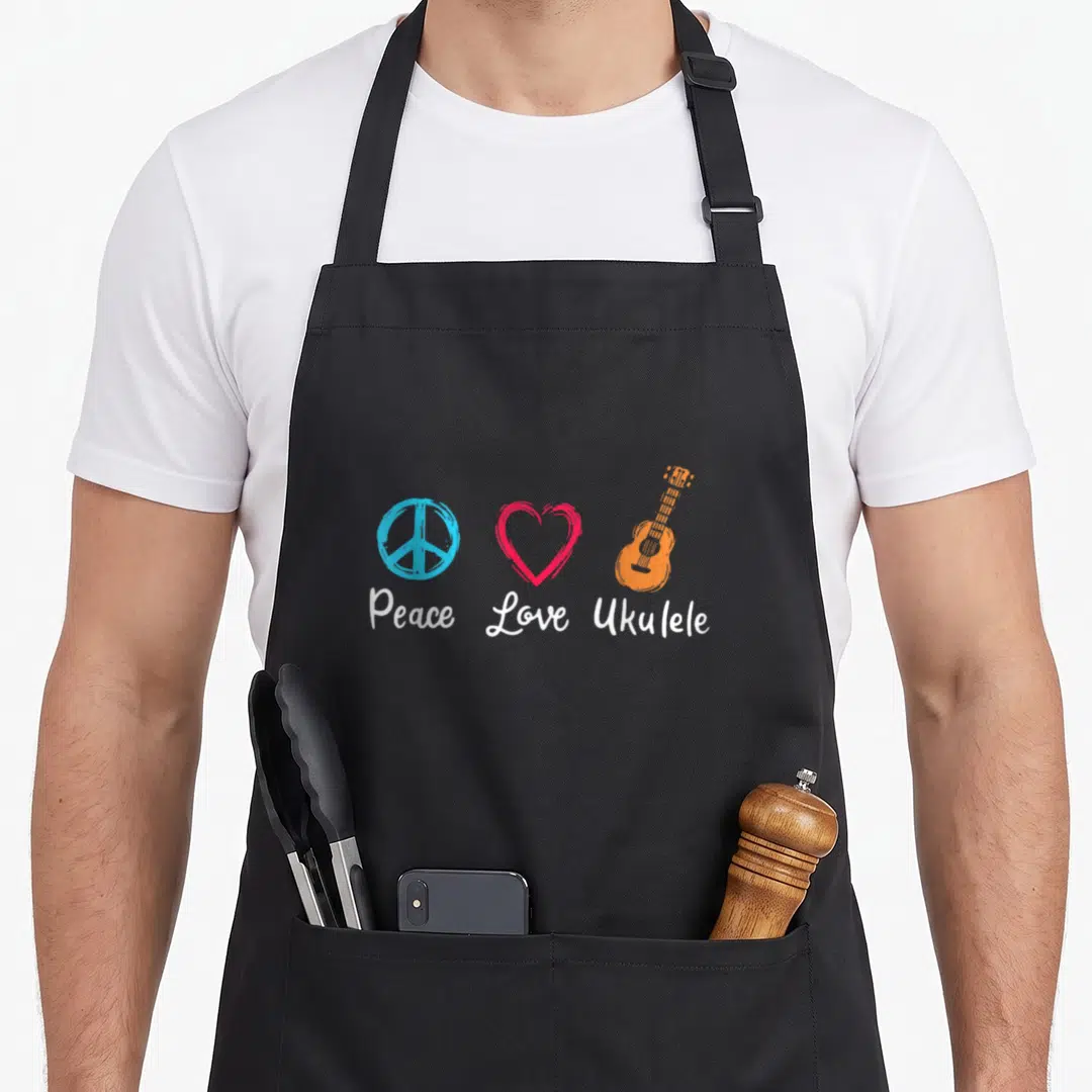 Peace ukulele apron - Three cartoon icons of a peace sign, heart, and ukulele in blue red and orange with white text — shown on a black cotton canvas apron against a white background