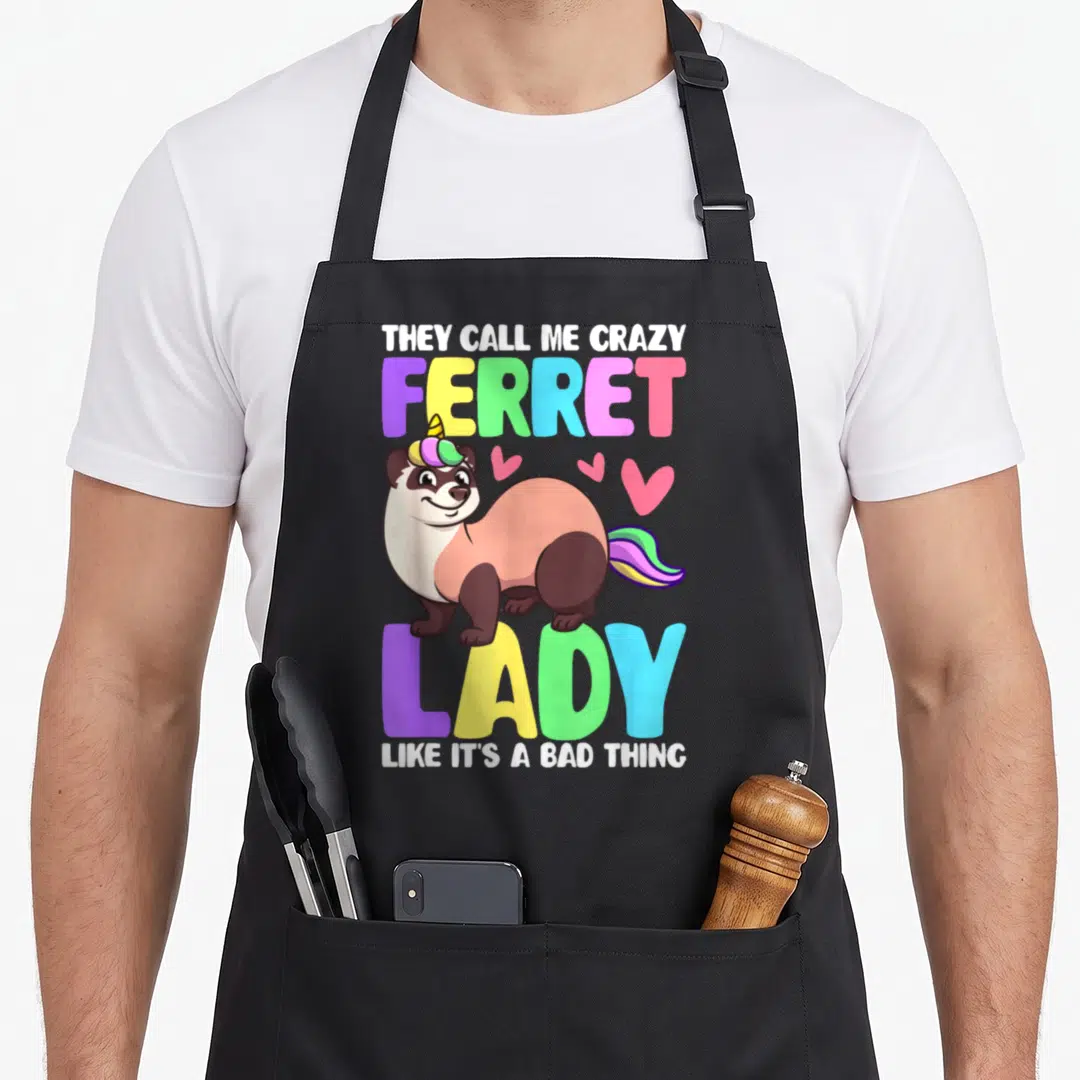 They crazy ferret lady apron - Cartoon ferret with rainbow horn and tail, colorful text, three pink hearts — shown on a black cotton canvas apron against a white background
