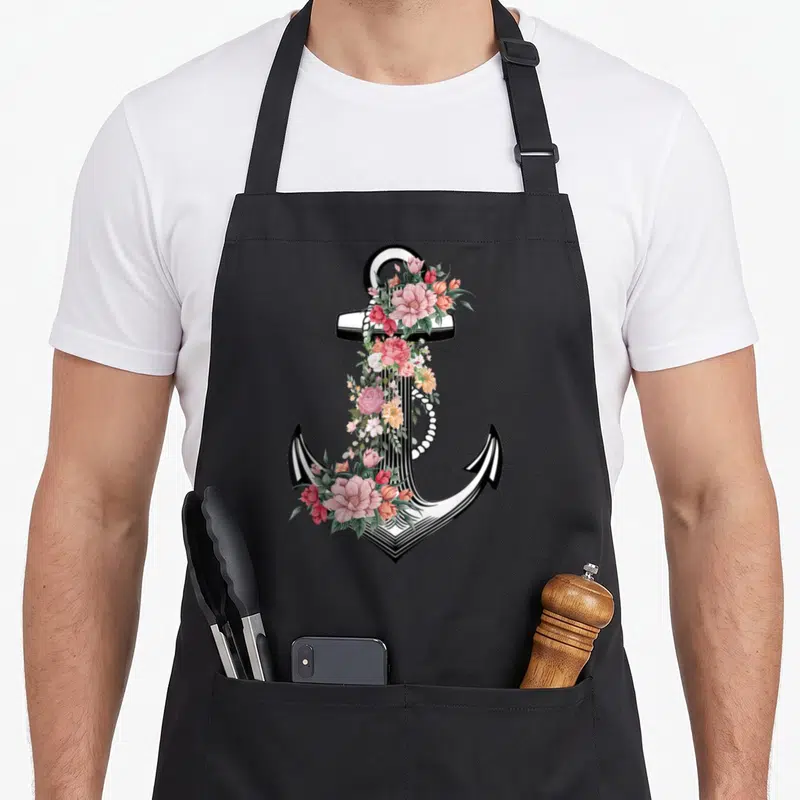 Boater nautical apron - White and black anchor with pink peach flowers and green leaves on black background — shown on a black cotton canvas apron against a white background