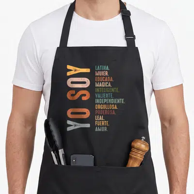 Soy hispanic aprons - Vertical YO SOY text next to stacked Latina adjectives in teal pink red yellow olive green light — shown on a black cotton canvas apron against a white background