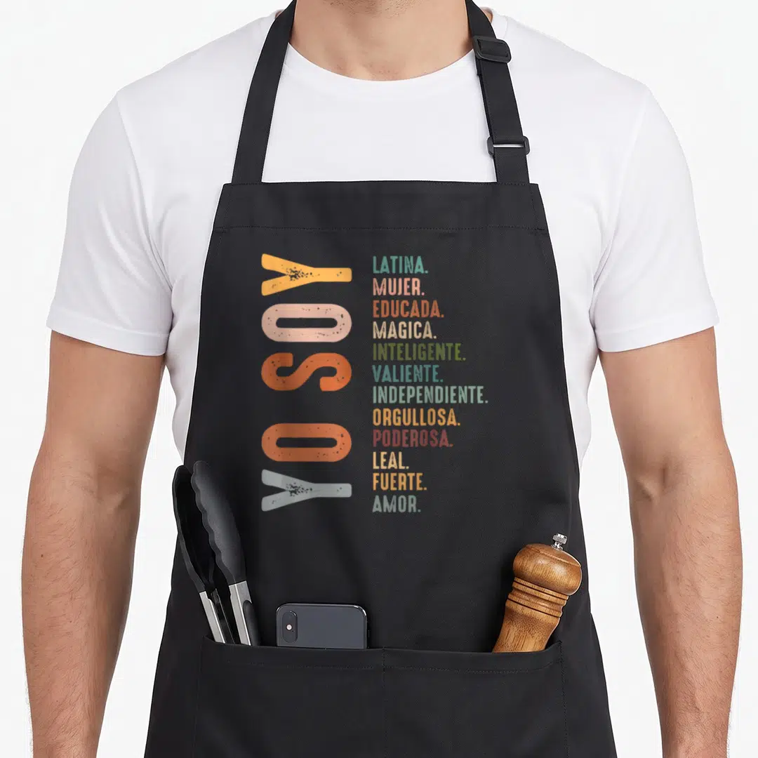 Soy hispanic aprons - Vertical YO SOY text next to stacked Latina adjectives in teal pink red yellow olive green light — shown on a black cotton canvas apron against a white background