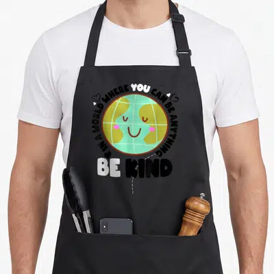 Bully unity day apron - Smiling cartoon earth with pink cheeks and wrapped text IN A WORLD WHERE YOU CAN BE ANYTHING — shown on a black cotton canvas apron against a white background