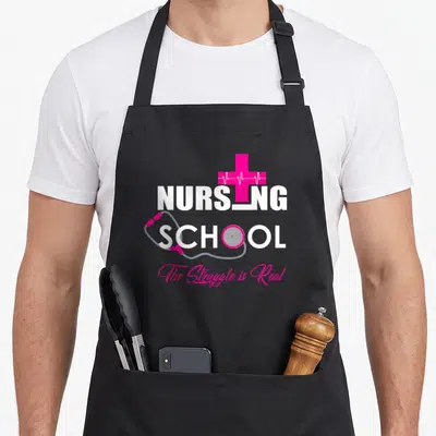 School nursing apron - Pink cross with white heartbeat line, grey stethoscope wrapping white NURSING SCHOOL text, pink — shown on a black cotton canvas apron against a white background