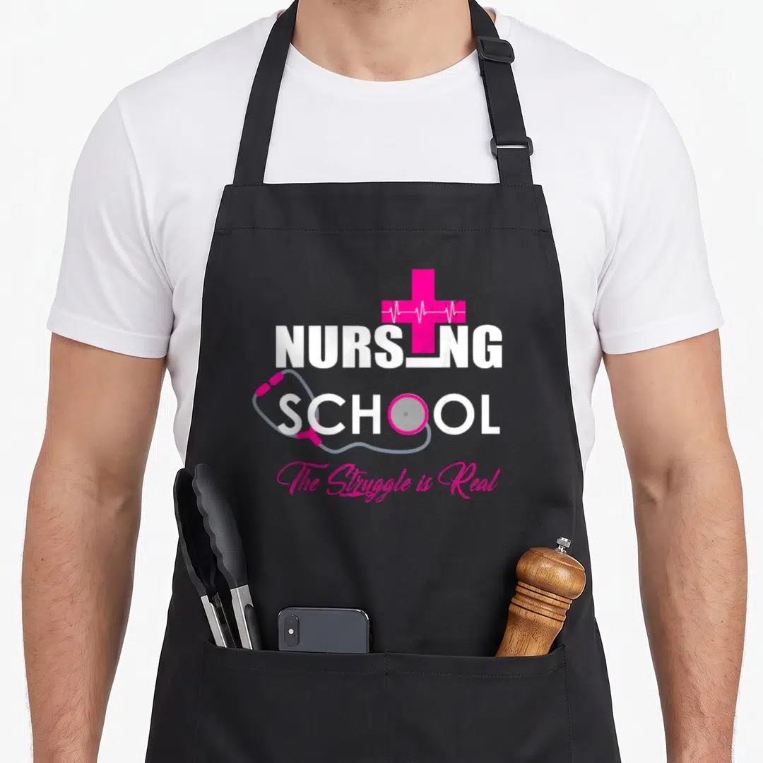 School nursing apron - Pink cross with white heartbeat line, grey stethoscope wrapping white NURSING SCHOOL text, pink — shown on a black cotton canvas apron against a white background