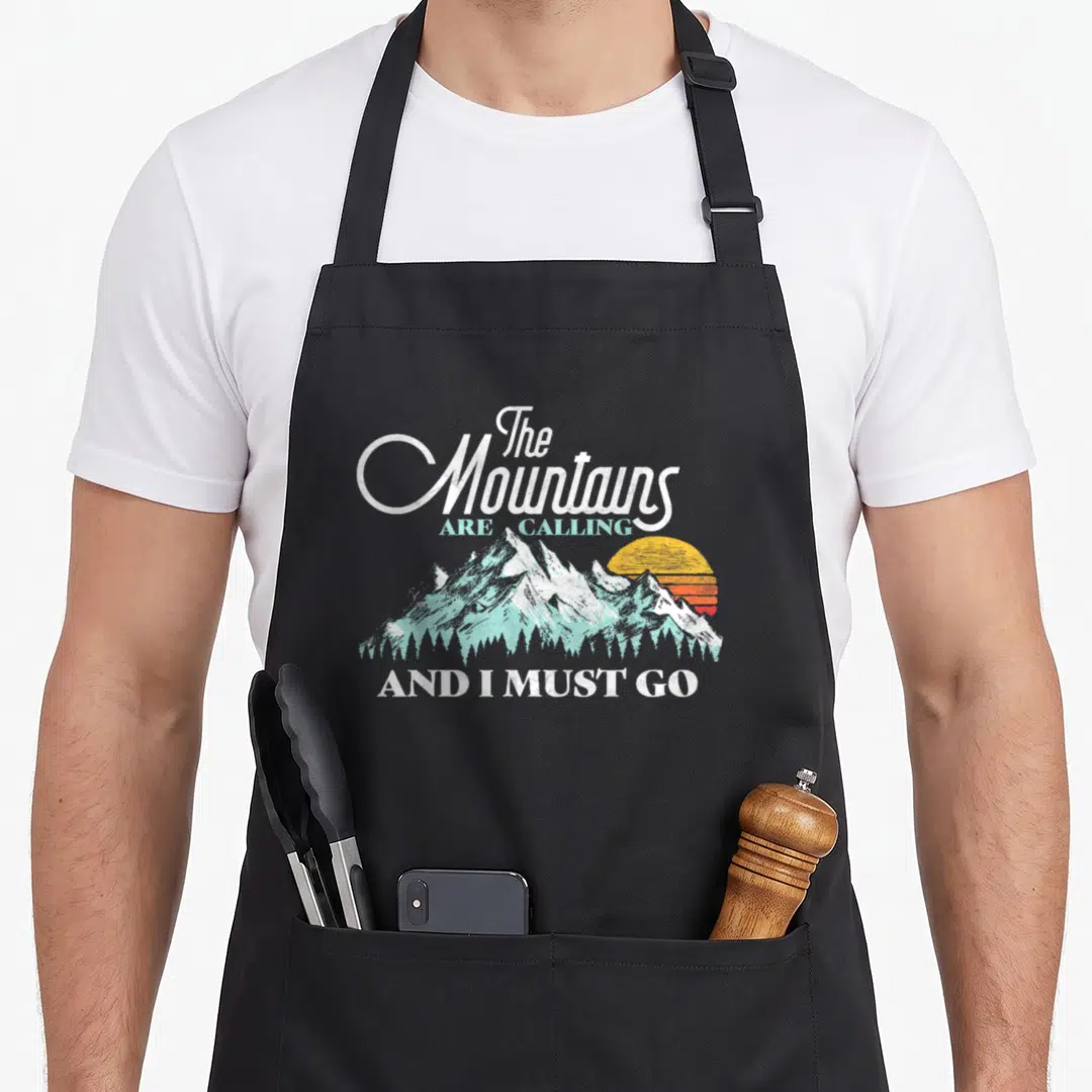Mountain 80s apron - Retro mountain illustration with sunset and trees surrounding the quote The Mountains ARE CALLING — shown on a black cotton canvas apron against a white background