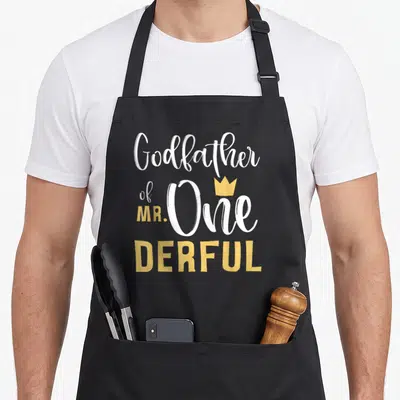 Godfather birthday apron - Stacked text Godfather of MR One Derful with small gold crown above the letter O — shown on a black cotton canvas apron against a white background