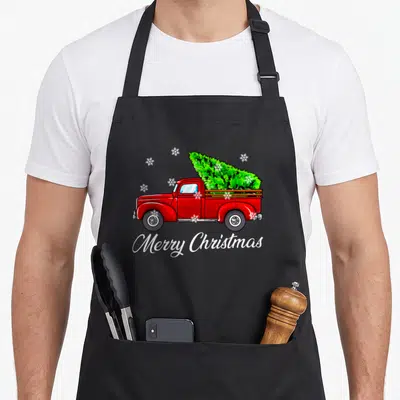 Truck christmas tree apron - Red truck carrying green pine tree with white Merry Christmas script and snowflakes — shown on a black cotton canvas apron against a white background