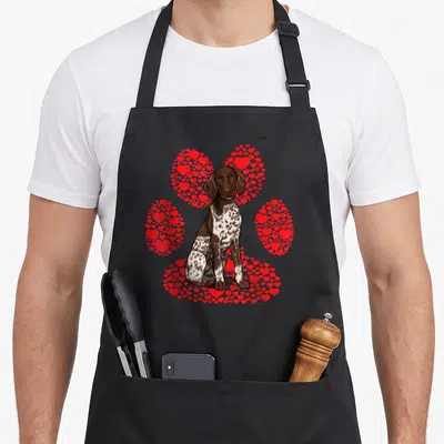 German valentine's day apron - Brown and white dog sitting in front of a paw print made of small red hearts — shown on a black cotton canvas apron against a white background