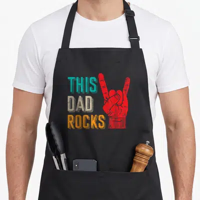 Desi music dad apron - Stacked This Dad Rocks text in teal beige orange next to red hand sign with distressed texture — shown on a black cotton canvas apron against a white background