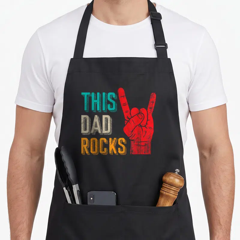 Desi music dad apron - Stacked This Dad Rocks text in teal beige orange next to red hand sign with distressed texture — shown on a black cotton canvas apron against a white background