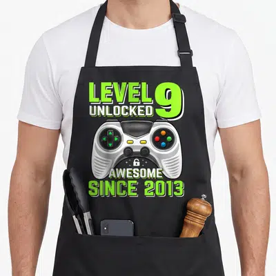 Level unlocked 9th birthday video game apron - Lime green text LEVEL 9 UNLOCKED above a grey game controller with SINCE — shown on a black cotton canvas apron against a white background