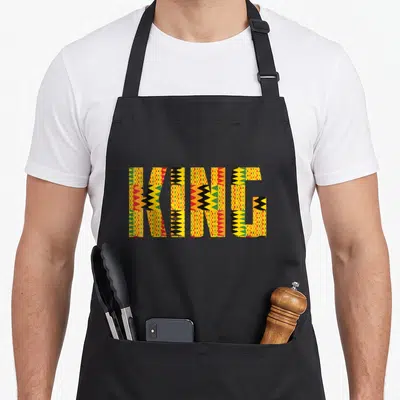 King african apron - KING text filled with yellow red green and black Kente cloth geometric patterns — shown on a black cotton canvas apron against a white background