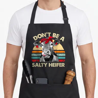 Salty farm apron - Realistic cow with red bandana and earrings under arching text Don't Be A Salty Heifer on retro — shown on a black cotton canvas apron against a white background