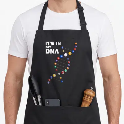 Dna billiards apron - Billiard balls arranged in a DNA helix shape next to stacked white text reading It's In My DNA — shown on a black cotton canvas apron against a white background