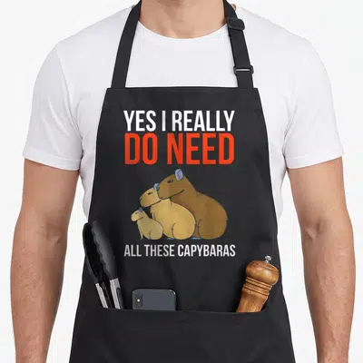 Capybaras do i need an apron - Three cartoon capybaras in brown tan and white below white and red text reading YES I — shown on a black cotton canvas apron against a white background