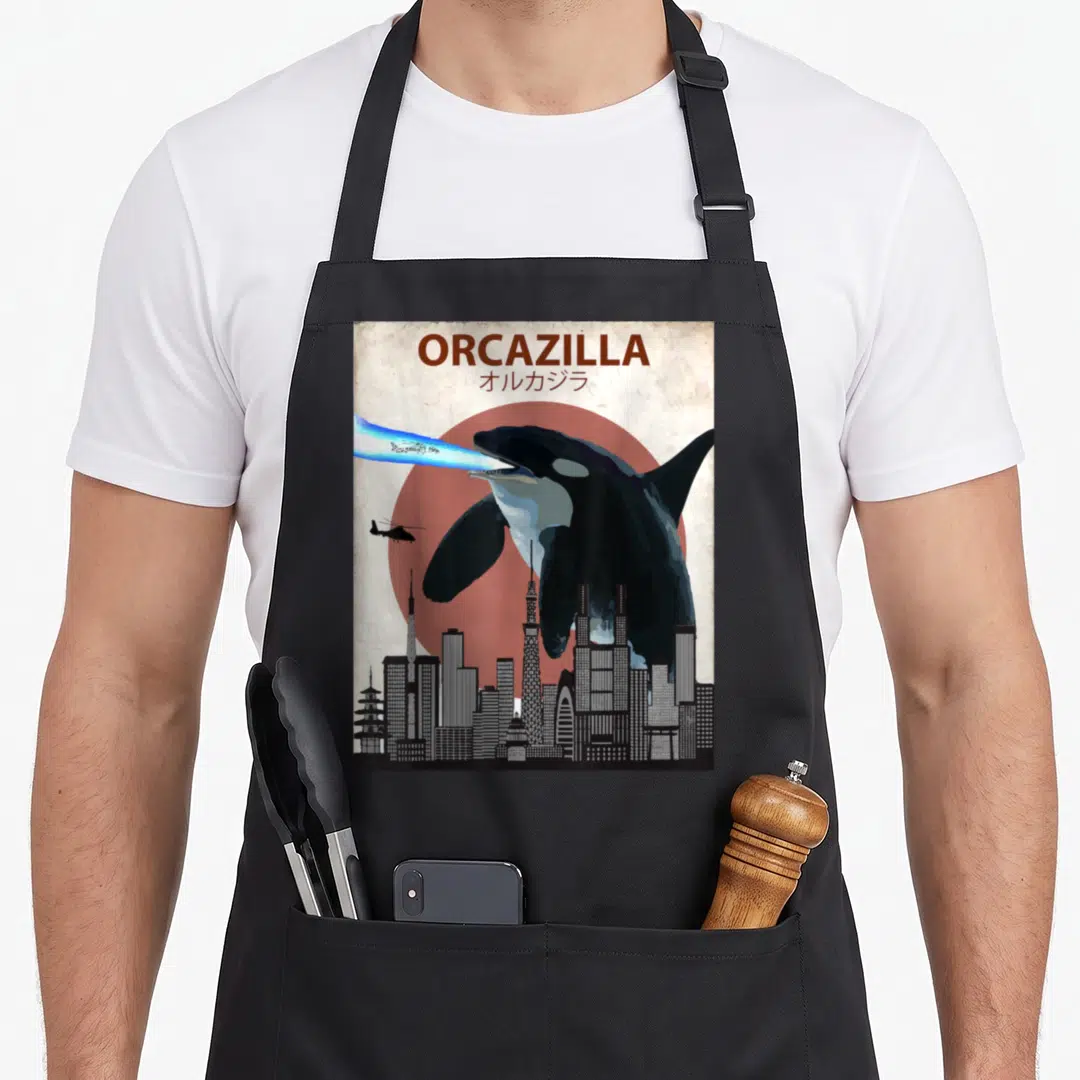 Killer orca dad apron - Retro poster of giant orca firing blue beam over city with ORCAZILLA text — shown on a black cotton canvas apron against a white background