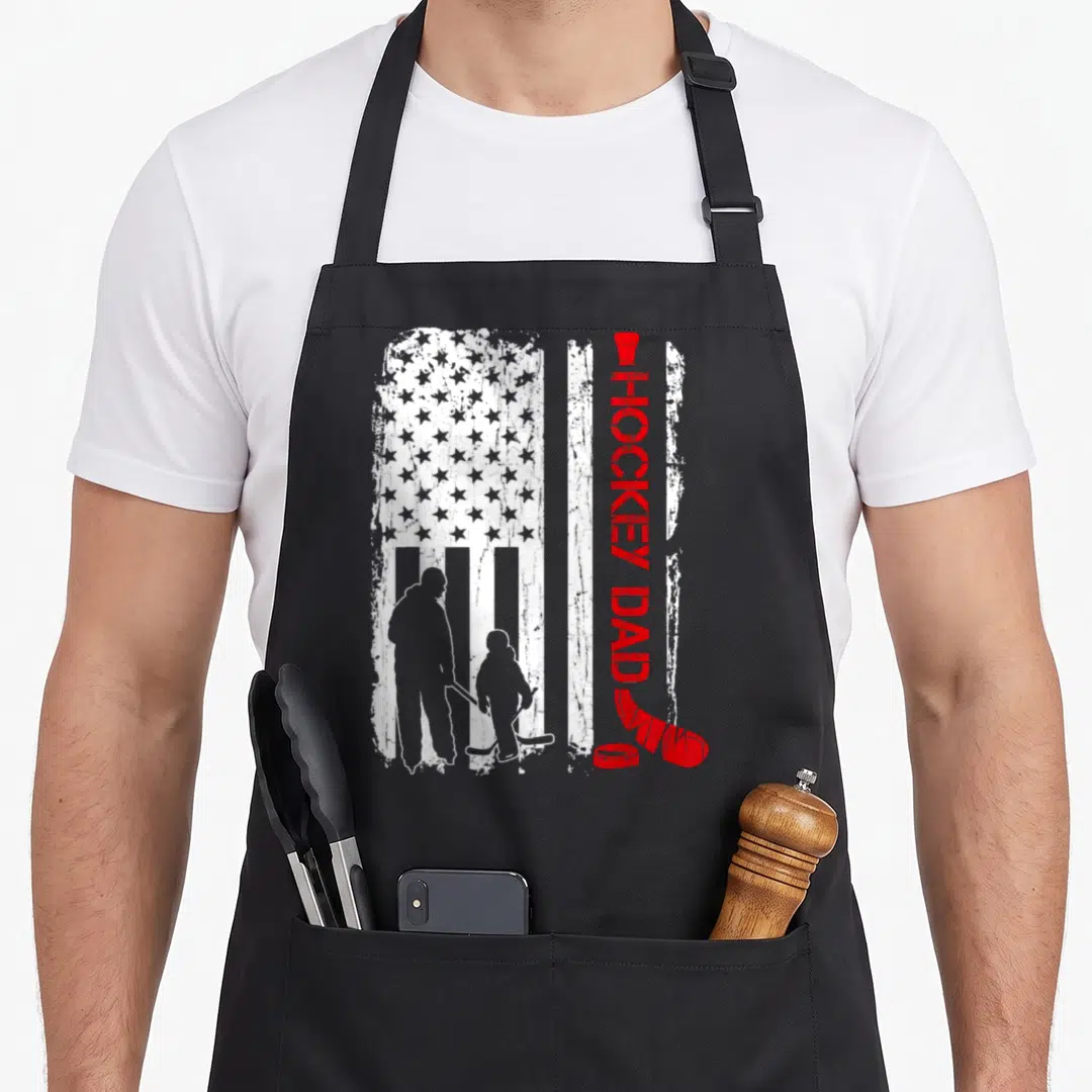 Funny hockey american flag apron - Vertical HOCKEY DAD text next to a father and child silhouette on a distressed flag — shown on a black cotton canvas apron against a white background