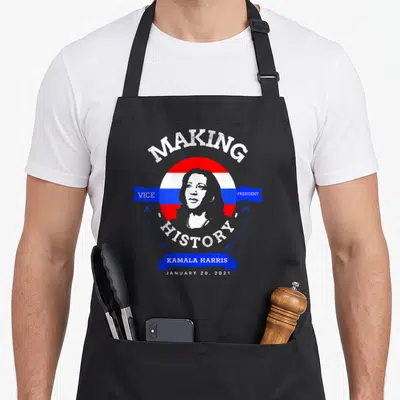 Kamala making aprons - Distressed portrait of Kamala Harris with Making Vice President History text on red white — shown on a black cotton canvas apron against a white background