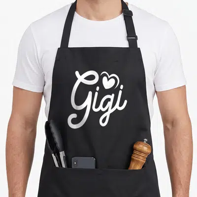 Grandchildren gigi apron - White script text Gigi with a heart symbol above the letter i — shown on a black cotton canvas apron against a white background