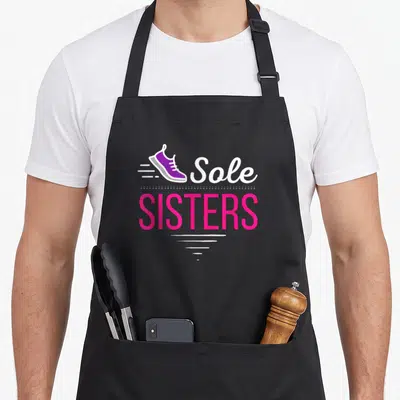 Sister running lover apron - Purple sneaker with Sole SISTERS text and pink lettering — shown on a black cotton canvas apron against a white background
