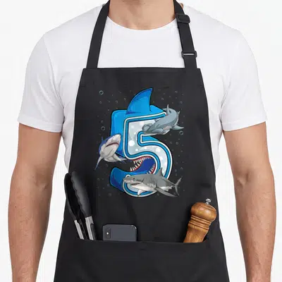 Ocean birthday apron - Large number 5 with three sharks and bubbles in blue, grey, and white cartoon art — shown on a black cotton canvas apron against a white background