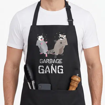 Garbage animal apron - Raccoon and opossum in sunglasses pointing at each other with GARBAGE GANG text below — shown on a black cotton canvas apron against a white background