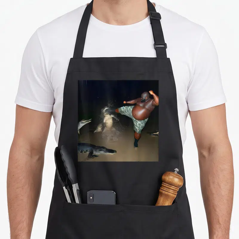 Kick meme apron - Large brown man in green shorts kicks pale alligator while dark crocodiles swim nearby in murky water — shown on a black cotton canvas apron against a white background