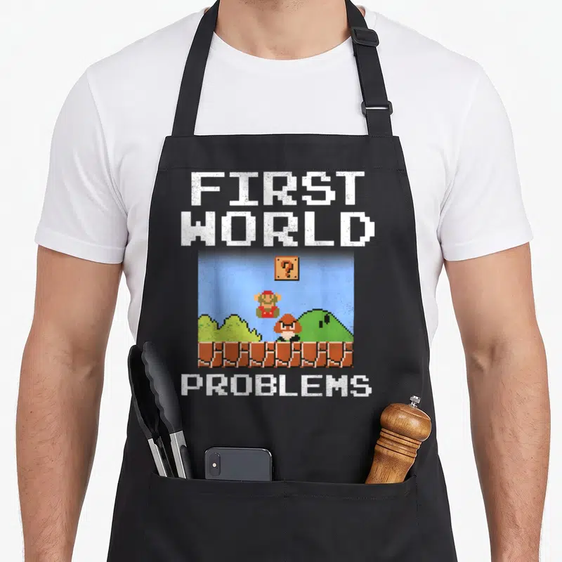 Bit novelty apron - Pixel art Mario and Toad on brick platform under question block with First World Problems text — shown on a black cotton canvas apron against a white background