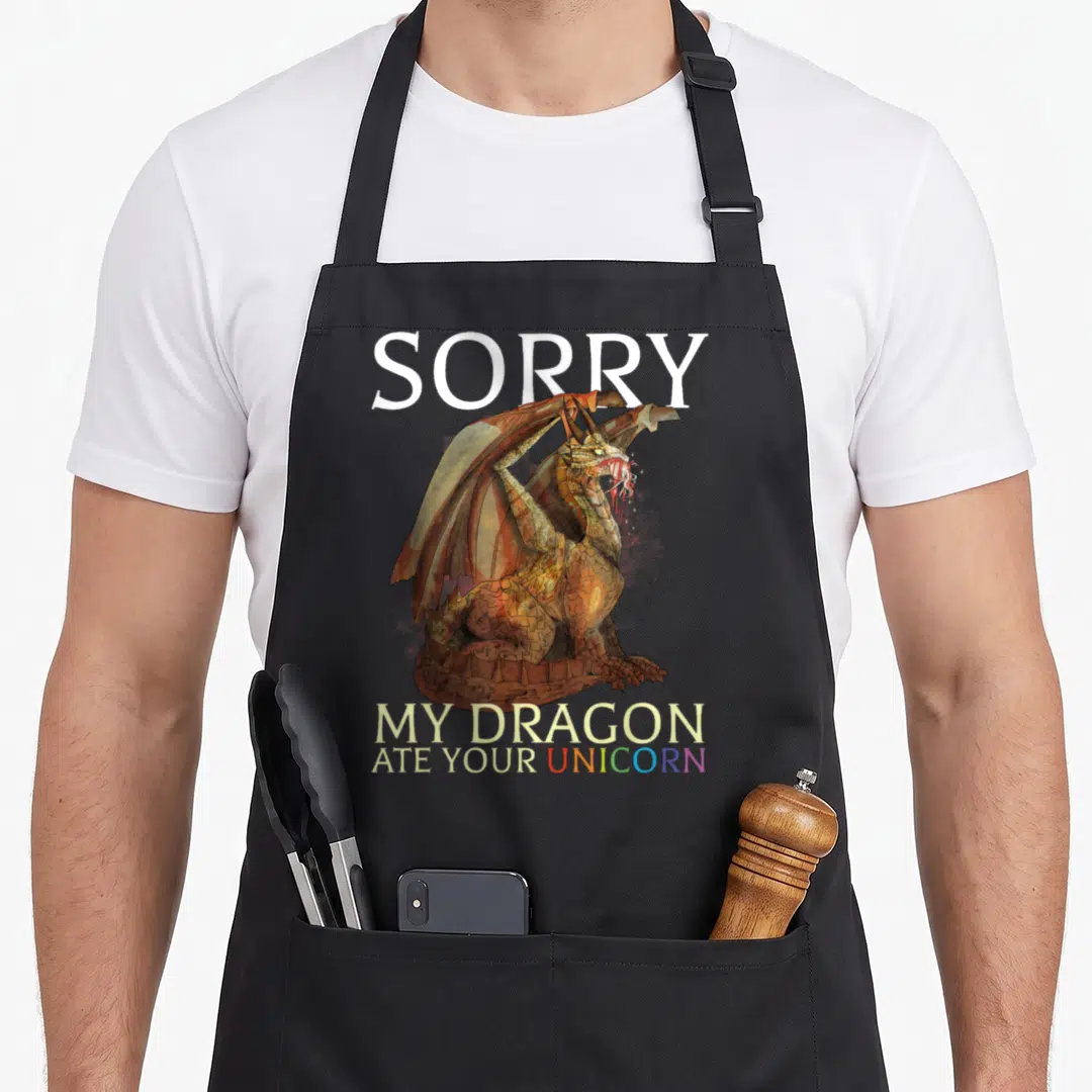 Ate dragon apron - Dragon sitting on rock holding unicorn horn, text SORRY MY DRAGON ATE YOUR UNICORN — shown on a black cotton canvas apron against a white background
