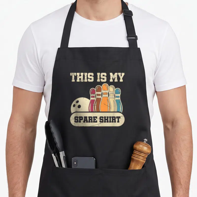 Spare gutter apron - Five colored bowling pins and ball below text THIS IS MY SPARE SHIRT on beige banner — shown on a black cotton canvas apron against a white background