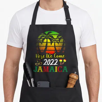Jamaica come and get it apron - Striped sun with palm trees, Here We Come 2022 Jamaica text, sunglasses, necklace — shown on a black cotton canvas apron against a white background
