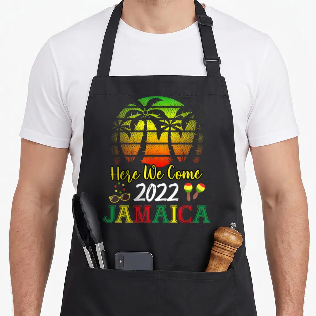 Jamaica come and get it apron - Striped sun with palm trees, Here We Come 2022 Jamaica text, sunglasses, necklace — shown on a black cotton canvas apron against a white background
