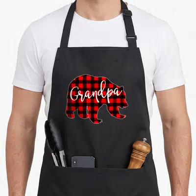 Grandpa buffalo plaid christmas apron - Red and black plaid bear silhouette with white Grandpa script text — shown on a black cotton canvas apron against a white background