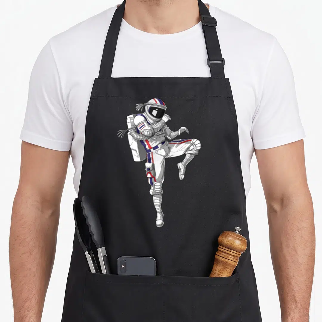 Muay boy astronaut apron - Cartoon astronaut in white suit with red blue stripes performing high kick with rope wraps — shown on a black cotton canvas apron against a white background