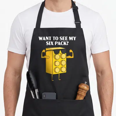See grill master apron - WANT TO SEE MY SIX PACK? text above yellow six-pack character with sunglasses flexing arms — shown on a black cotton canvas apron against a white background