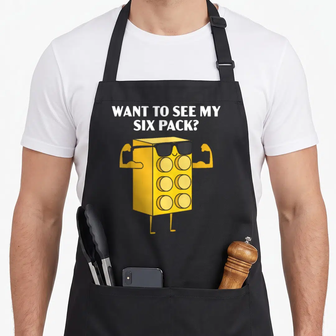 See grill master apron - WANT TO SEE MY SIX PACK? text above yellow six-pack character with sunglasses flexing arms — shown on a black cotton canvas apron against a white background