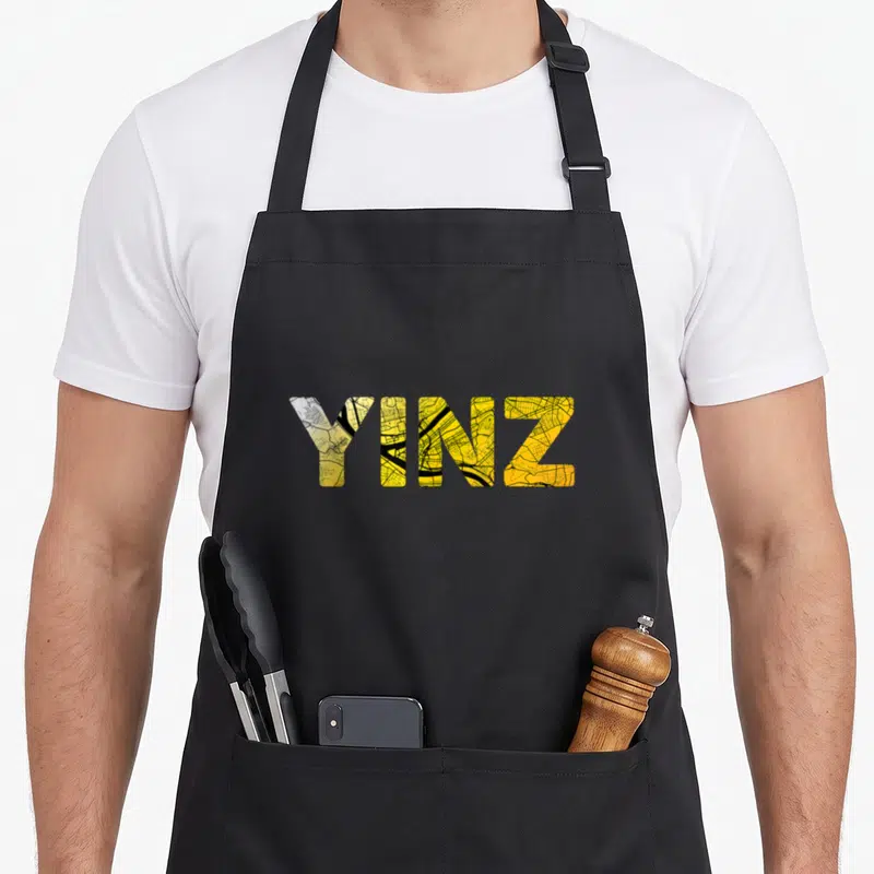 Pittsburgh pollia apron - YINZ text filled with yellow and black street map lines — shown on a black cotton canvas apron against a white background