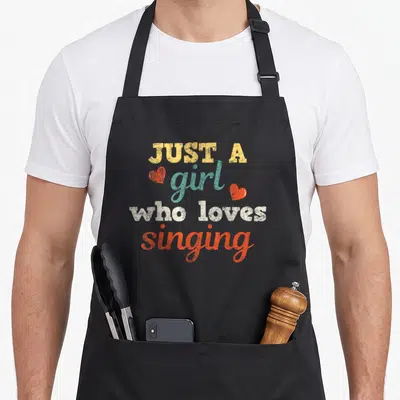 Music singing apron - Stacked text reading Just A Girl Who Loves Singing with two orange hearts on yellow teal white — shown on a black cotton canvas apron against a white background