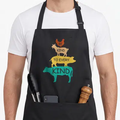 Kind vegetarian vegan apron - Stacked chicken goat pig cow silhouettes in orange red tan yellow teal with text inside — shown on a black cotton canvas apron against a white background