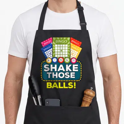 Shake bingo apron - Five colorful bingo cards above six lettered balls and a marquee sign reading SHAKE THOSE BALLS! — shown on a black cotton canvas apron against a white background