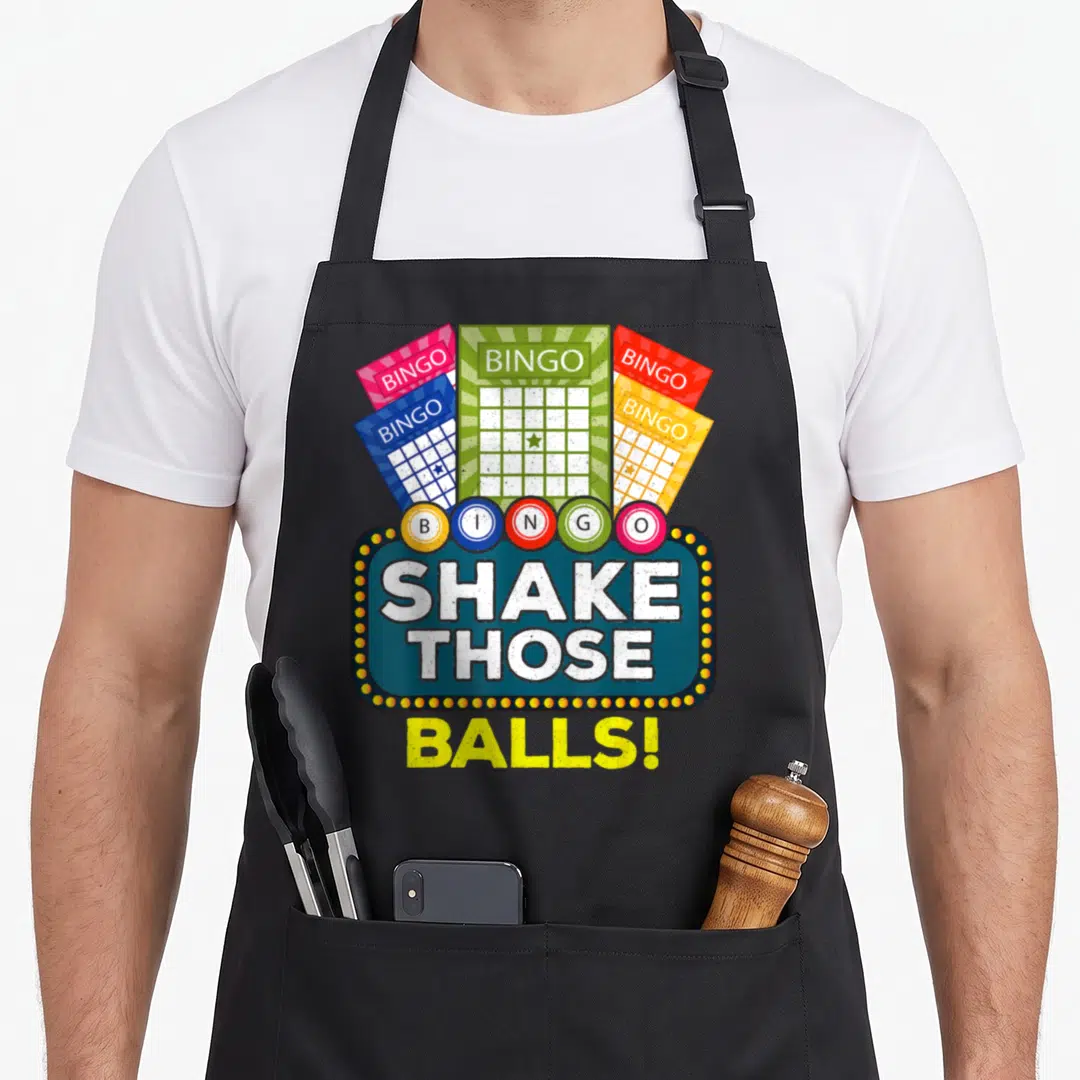 Shake bingo apron - Five colorful bingo cards above six lettered balls and a marquee sign reading SHAKE THOSE BALLS! — shown on a black cotton canvas apron against a white background