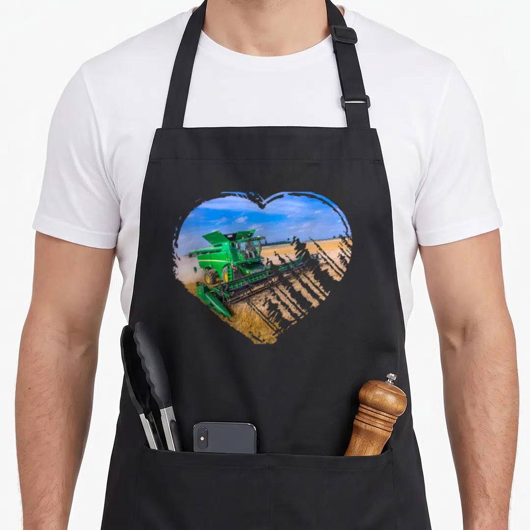 Funny combine harvester apron - Green combine harvester in a field cropped into a heart shape against a blue sky — shown on a black cotton canvas apron against a white background