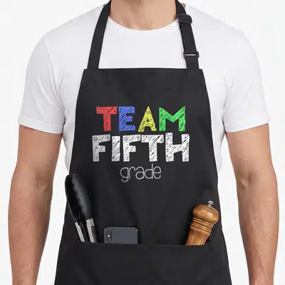 5th fifth grade for teacher student apron - Stacked text reading TEAM FIFTH grade in red blue yellow green and white — shown on a black cotton canvas apron against a white background